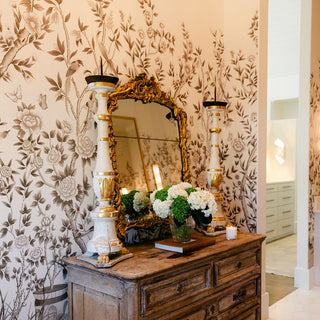 Amy Howards Cherbourg Chinoiserie Wall Mural from their 2025 wallpaper collection featured in a master bathroom with a beautiful aged antique chest of drawers in front of it. 