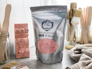A package of Toscana Milk Paint in pink color, displayed alongside brushes and a painted decorative item, with tools and a painted surface in the background.
