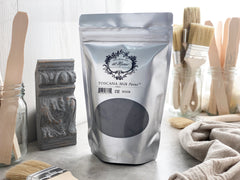 A package of Toscana Milk Paint in color Noir Black, displayed alongside brushes and a piece of furniture for restoration.