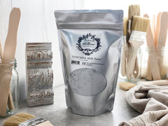 A package of Toscana Milk Paint in Scandinavian Grey color, displayed alongside brushes and a piece of furniture being refurbished.