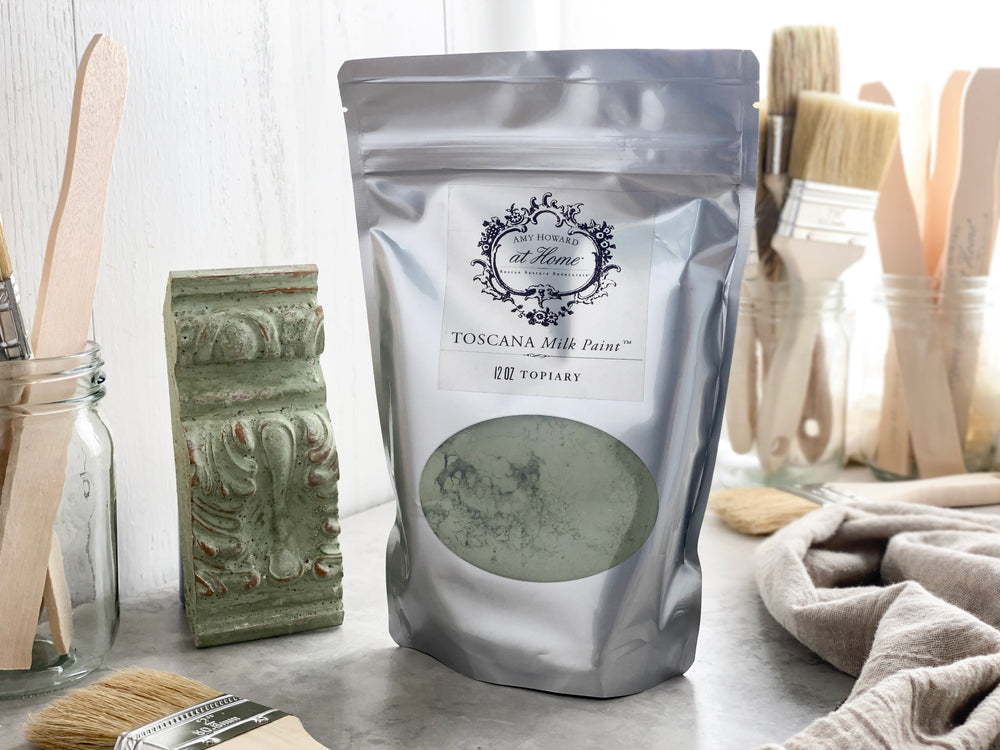 A package of Toscana Milk Paint in 'Topiary' color, with a background featuring brushes and a decorative ornament.