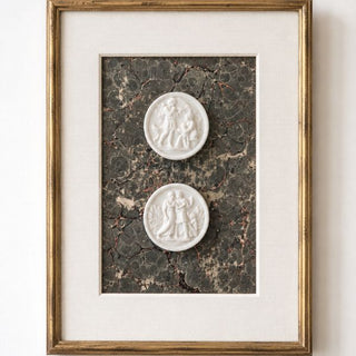 Two CeramiCast classical figure medallion castings from the Amy Howard At Home Intaglio Decorative Mould displayed as framed wall art in an antique gold gilt frame with linen mat and antique marbled paper backing on a white wall