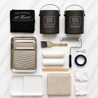 Amy Howard At Home Manor Borne Wall Paint curated collection kit contents including two gallons of Manor Borne Ultra-Scrub Matte paint, brush, roller covers, roller frame, roller pan, drop cloth, painter's tape, stir stick, rags, and branded apron on marble background