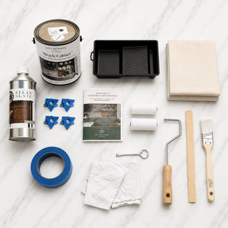 Amy Howard At Home Miracle Kitchen Cabinet Kit contents including Miracle Cabinet Paint gallon, Clean Slate degreaser, roller pan, canvas drop cloth, blue painter's tape, paint stands, roller covers, wooden handle roller, nylon brush, can opener, stir stick, rags, and step-by-step guide on marble background