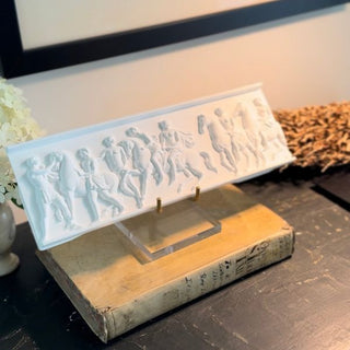 CeramiCast classical frieze panel casting from the Amy Howard At Home Relief Decorative Mould displayed on an acrylic and brass stand on a dark surface with antique book and white hydrangeas