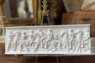 CeramiCast classical frieze panel casting from the Amy Howard At Home Relief Decorative Mould displayed on an acrylic and brass stand on a dark surface with antique book and white hydrangeas