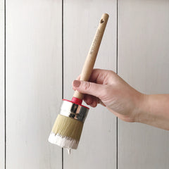 A hand holding a round synthetic hog hair brush with a wooden handle great for waxing furniture paint brush